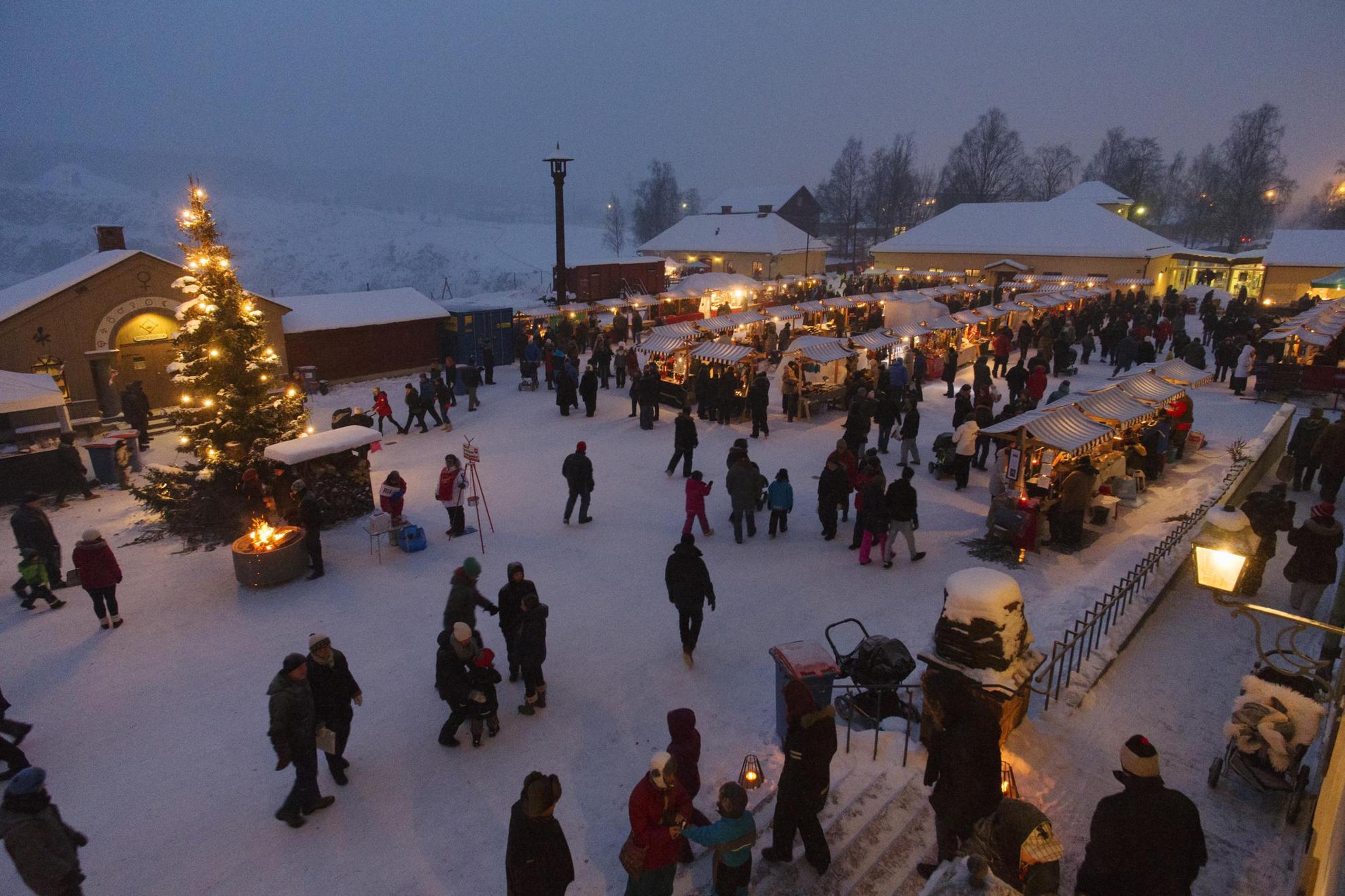 Un marché de Noël sous la neige avec de nomnbreuses étals, des personnes et un grand sapin de Noël à côté d'un feu.