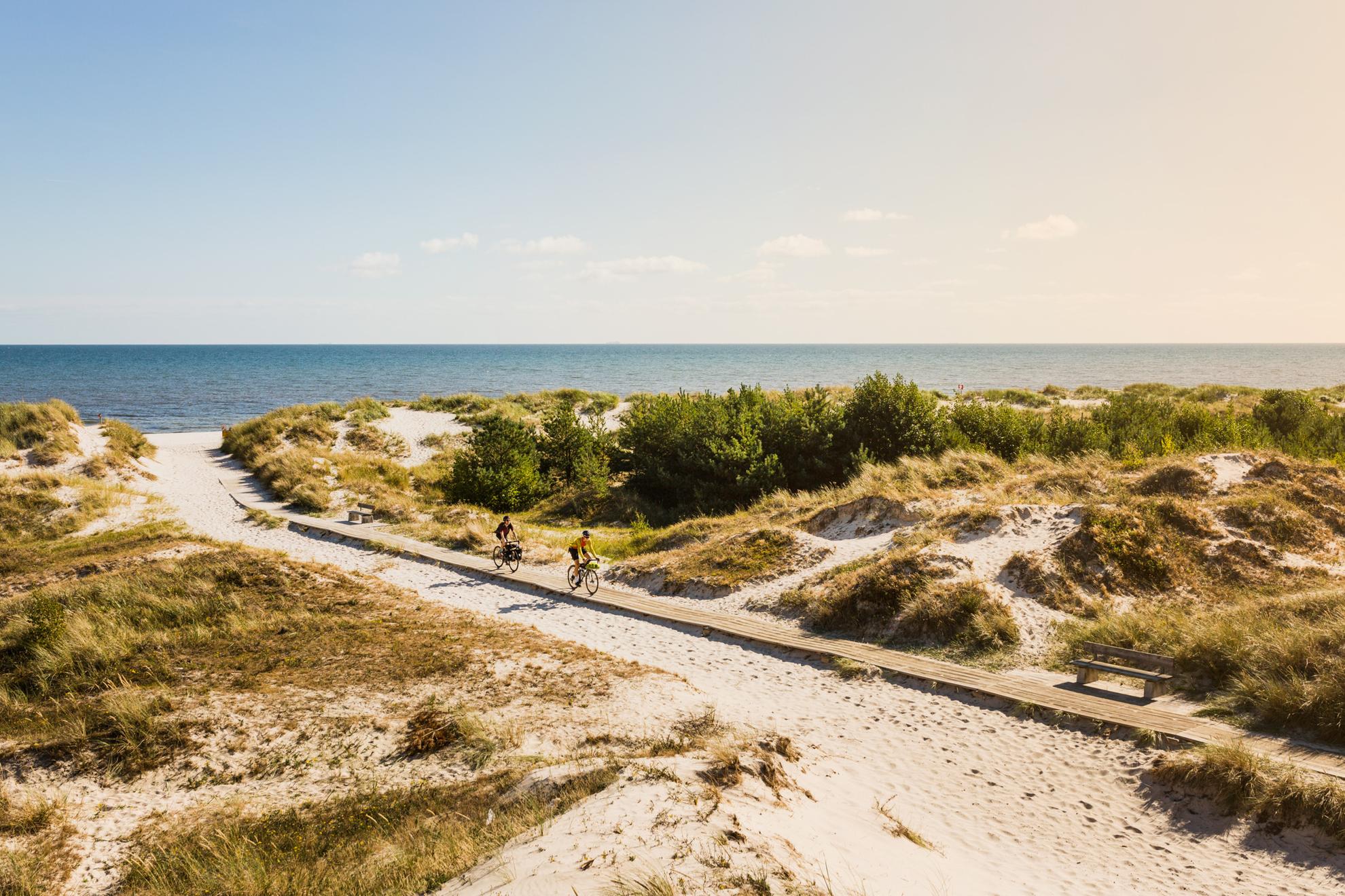 Deux personnes font du vélo sur une passerelle en bois sur une plage de sable entourée de touffes d'herbe.