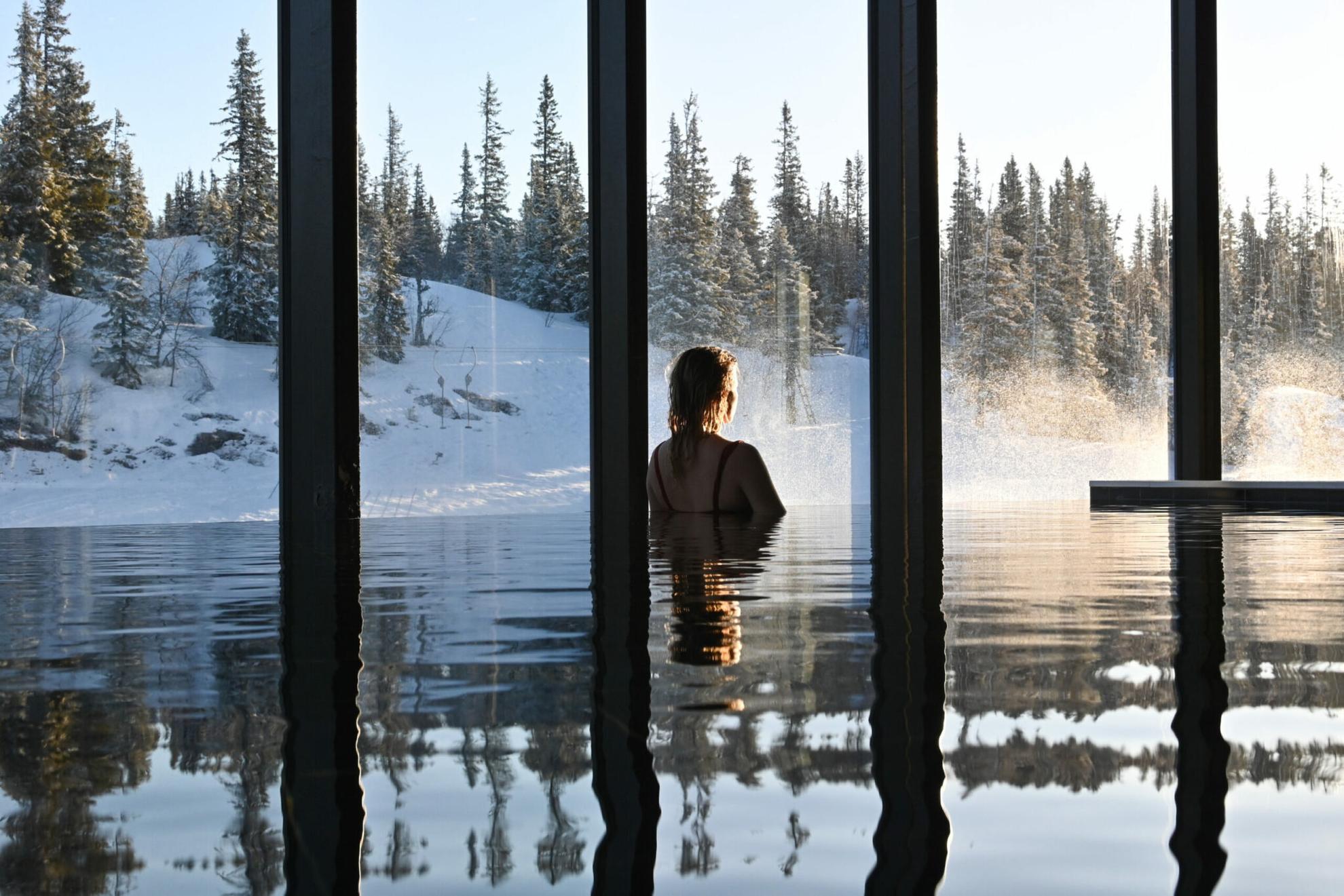 Une personne dans une piscine intérieure à débordement regardant la forêt enneigée à travers les grandes fenêtres.