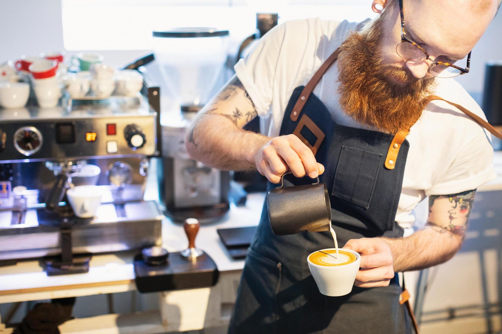 A man wearing a white t-shirt, glasses and a thick beard and an apron is pouring steamed milk into an espresso cup.