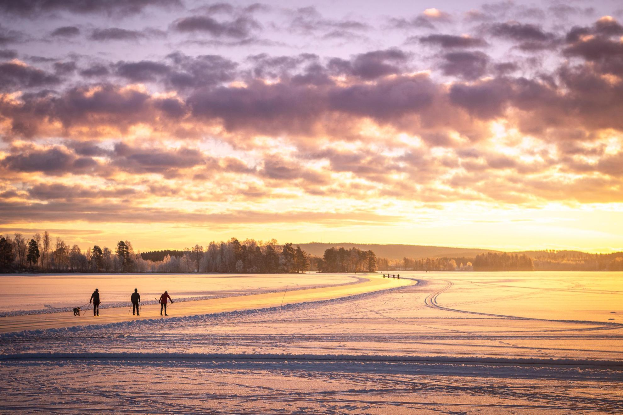 Silhouette de trois personnes en train de patiner sur un lac gelé. Le soleil brille d'une lueur orange.