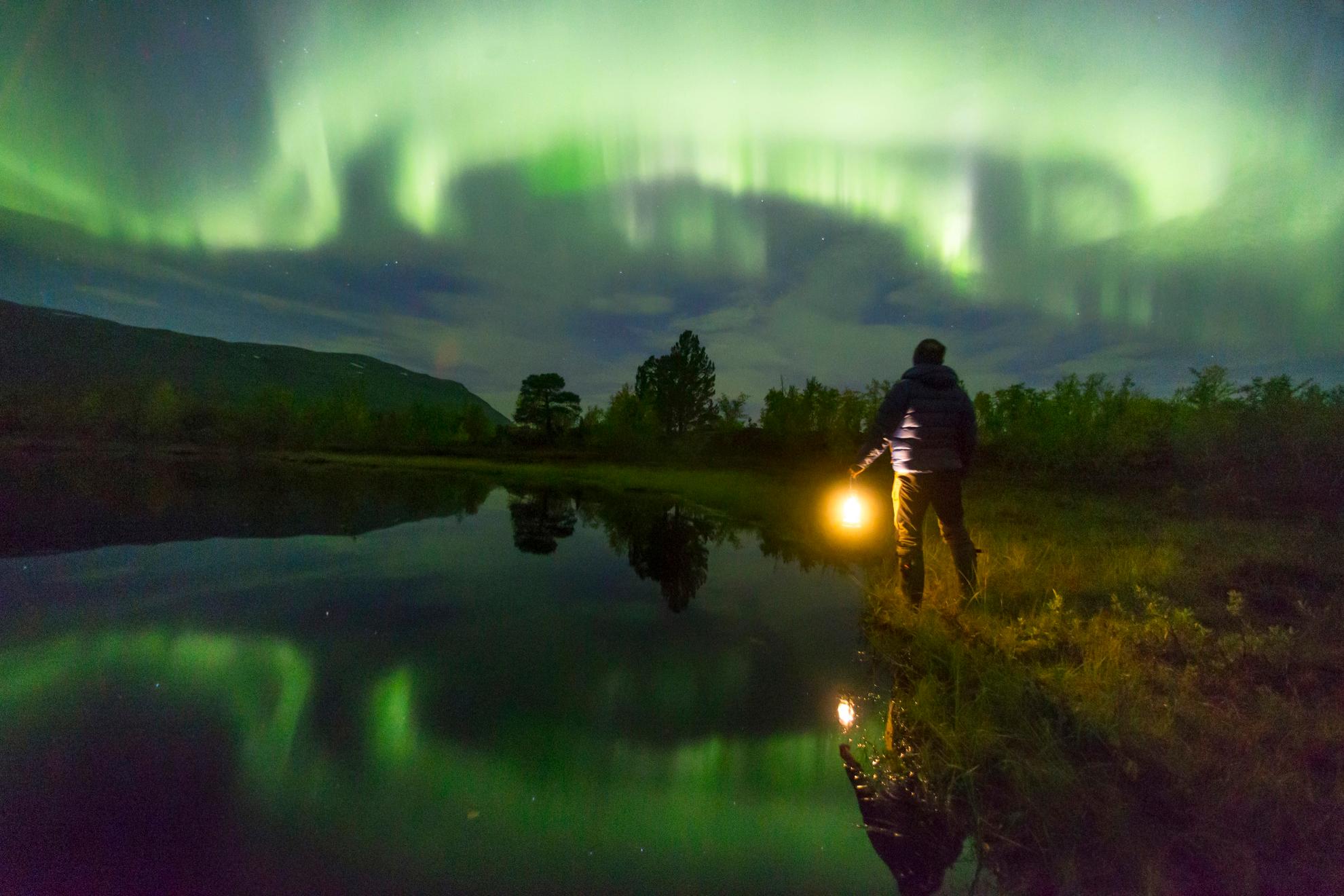 Une personne tenant une lampe regarde les aurores boréales vertes dans le ciel. Les aurores boréales se reflètent dans le lac à côté de la personne.