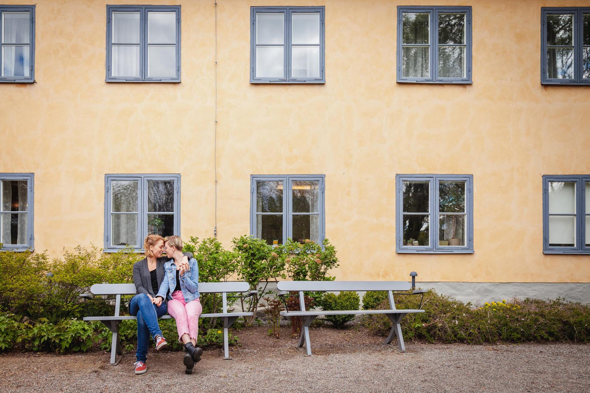 Deux femmes s'embrassant et se tenant la main, assises sur un banc devant une maison.
