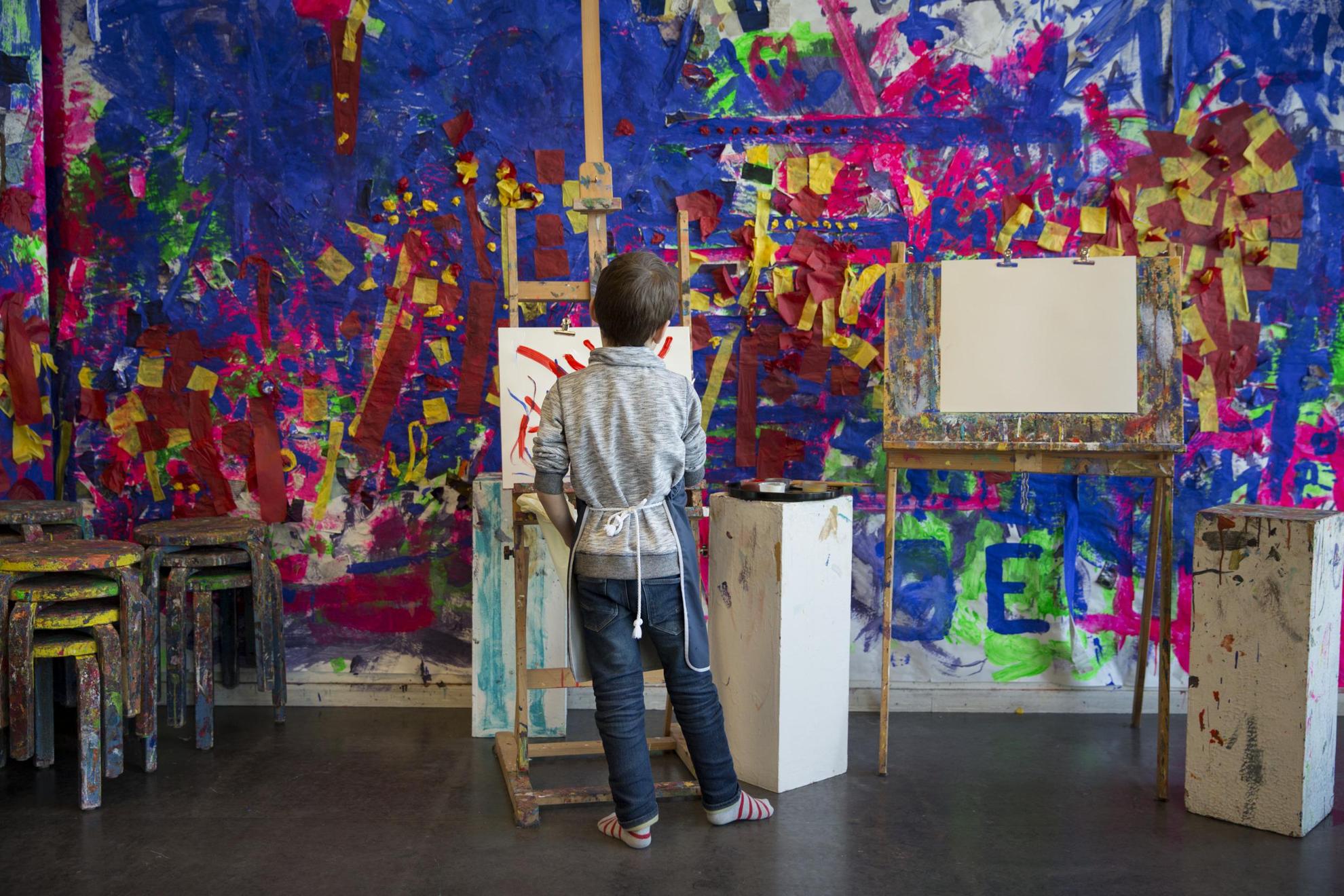 A "Room for Children", un jeune garçon peint sur une toile posée sur un chevalet. Les murs en arrière-plan et les chaises sont recouverts de peinture.