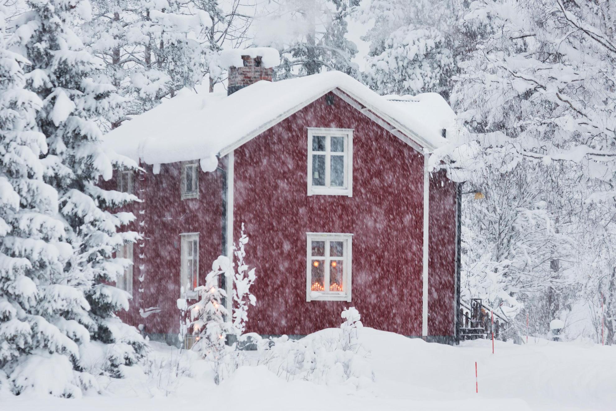 Une maison en bois rouge avec des encadrements blancs est couverte de neige et il neige en parallèle.