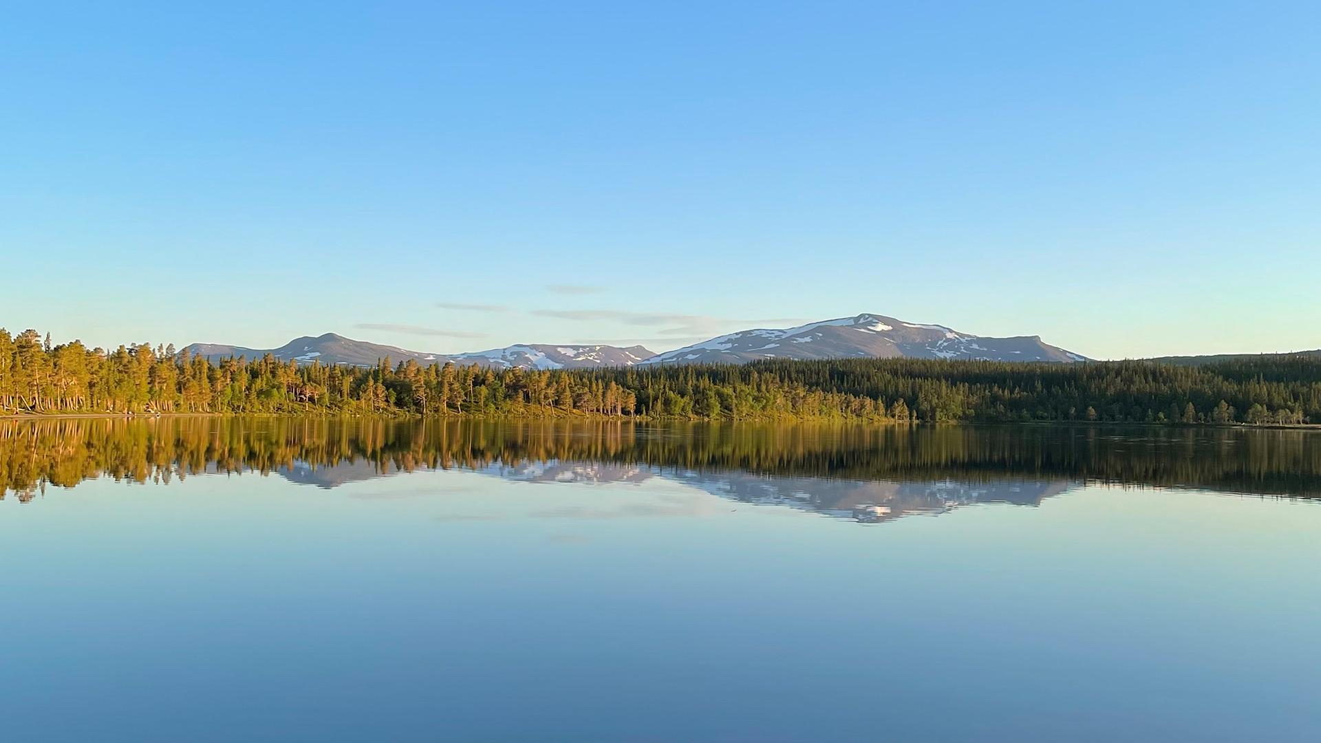L'eau du lac Nulltjärn est calme et reflète le ciel bleu, les forêts et les montagnes environnantes.
