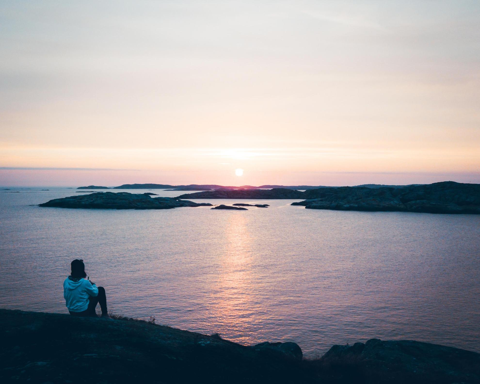 Une personne assise sur une falaise au dessus de la mer et qui regarde le coucher du soleil.